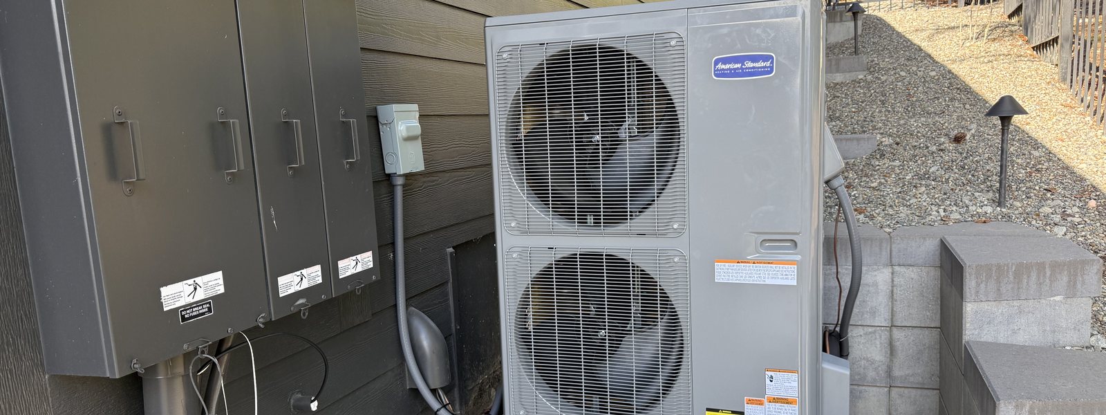 American Standard outdoor unit beside a home's electrical panel next to a gravel side yard