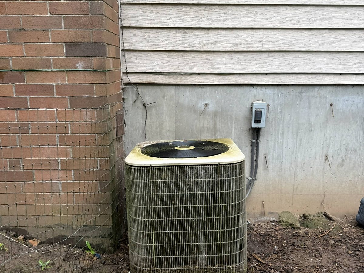 Older outdoor condenser with a weathered top beside a brick wall